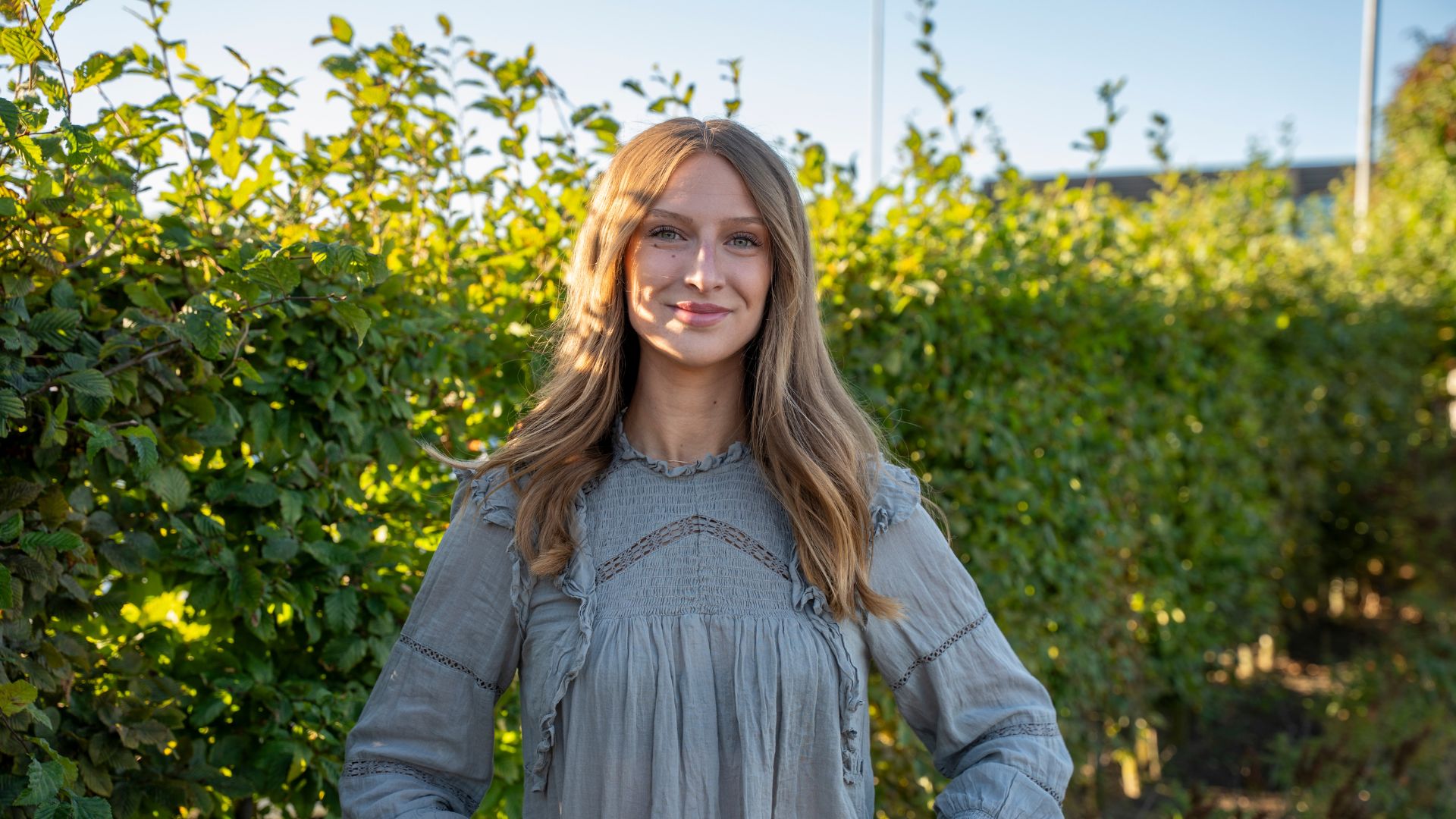 Porträtt på Wendla, projektledare och affärsutvecklare på Sandberg Development, framför en grön häck i solnedgången.